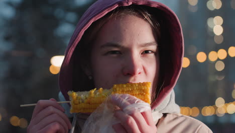 mujer joven comiendo maíz en un palo envuelto en plástico blanco, vapor que se eleva desde el maíz fresco, luces bokeh en un fondo borroso, disfrutando de un bocadillo al aire libre en la noche de invierno con luces cálidas