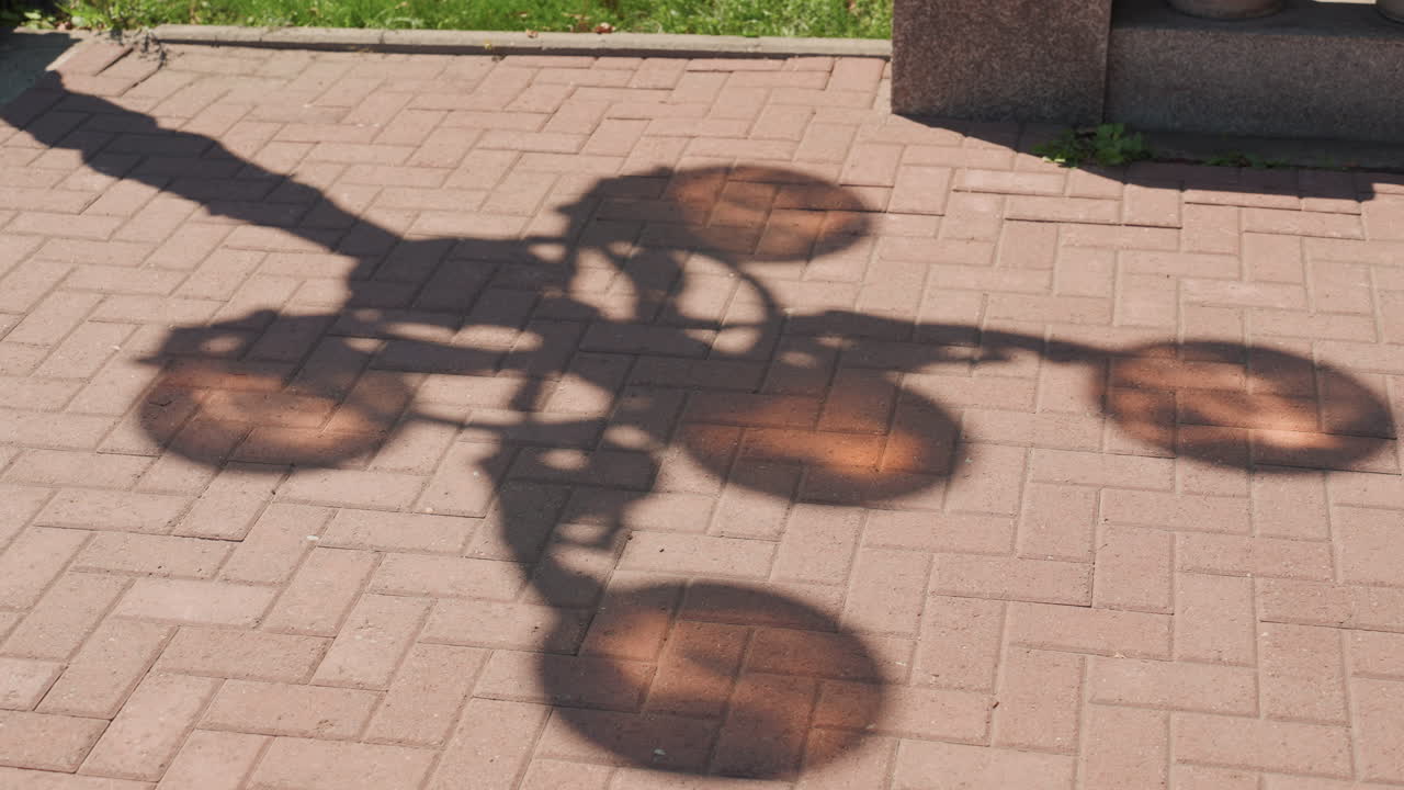 sunlit ornate lamppost shadow on brick paving, warm amber pools from round lanterns, decorative metalwork silhouette stretching across textured pavers, late afternoon golden hour glow, park border