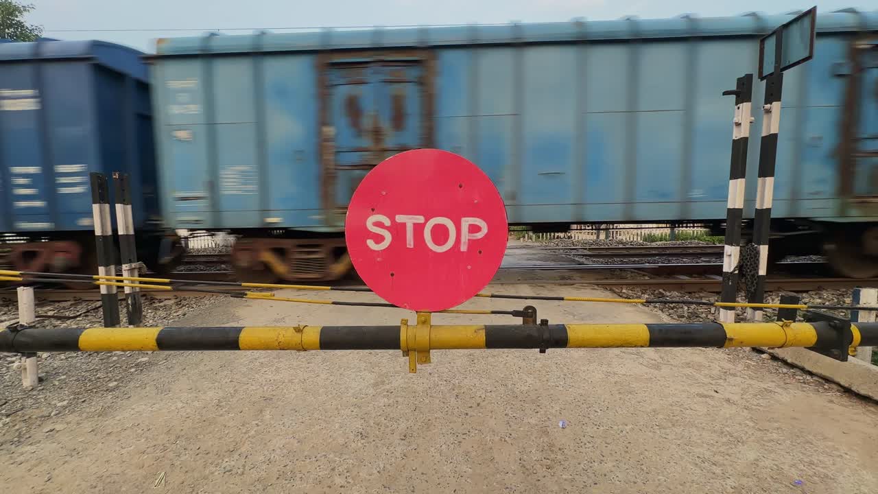 Blue freight train rushes past a rural railway crossing as the yellow barrier with a red STOP sign blocks the gravel road, creating a fast moving backdrop