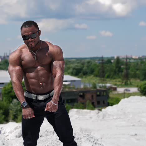 Male bodybuilder flexing muscles. African american sportsman standing shirtless showing muscles outdoors. Dark-skinned athlete in black glasses shows strong arms