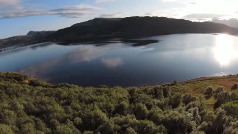 Flying-along-a-mountain-down-to-the-lake-overlooking-the-grassy-landscape-and-many-trees-on-the-hillside,-then-flying-over-the-lake-with-its-smooth-surface