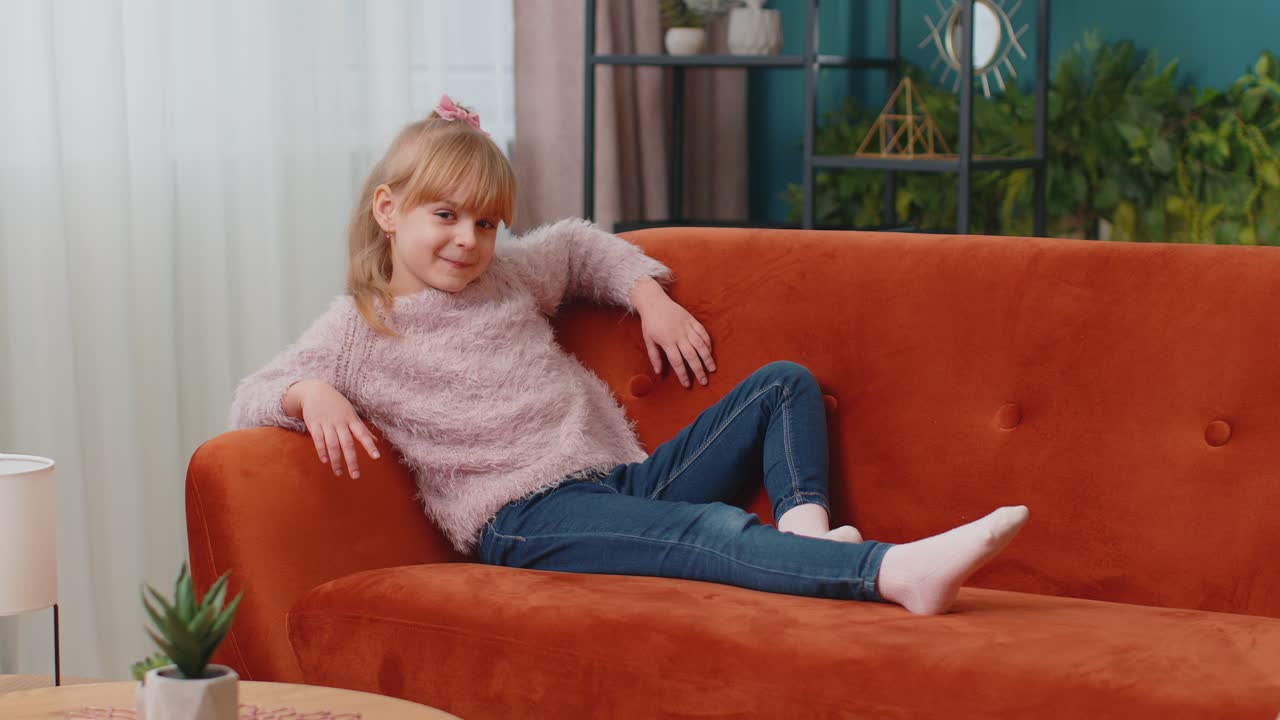 Child girl lying on sofa at home looking at camera smiling waving hands gesturing hello or goodbye