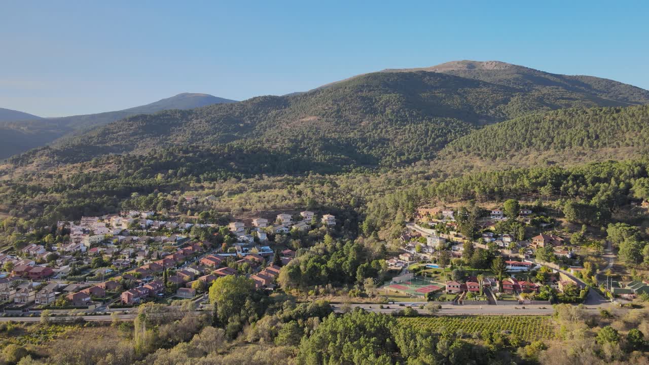 Riprese con drone di aree residenziali in collina con chalet a Santa María del Tiétar. Una strada dritta scorre sotto con veicoli in movimento, circondata da foreste verdi e montagne sotto un cielo blu.