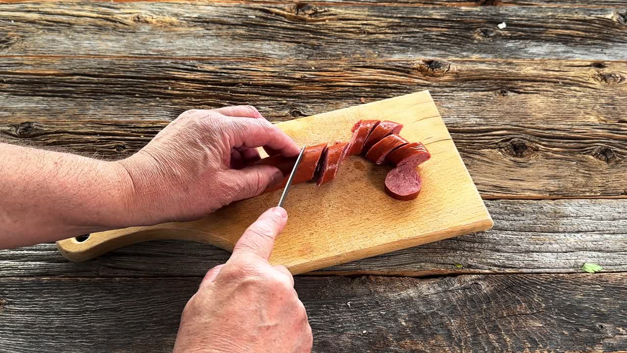 Mani maschili che usano un coltello affilato per affettare una tradizionale salsiccia affumicata su un tagliere rustico di legno. Vista dall'alto della preparazione della carne.