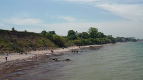 Kleiner-Abgelegener-Ostseestrand-An-Grünen-Bäumen-In-Brodten,-Deutschland,-Touristen,-Die-Sonnigen-Tag-Genießen,-Luftdrohnenansicht,-Die-Vorwärts-über-Meerwasser-Fliegt,-Erhebt-Sich-Und-Enthüllt-Ein-Riesiges-Grünes-Feld