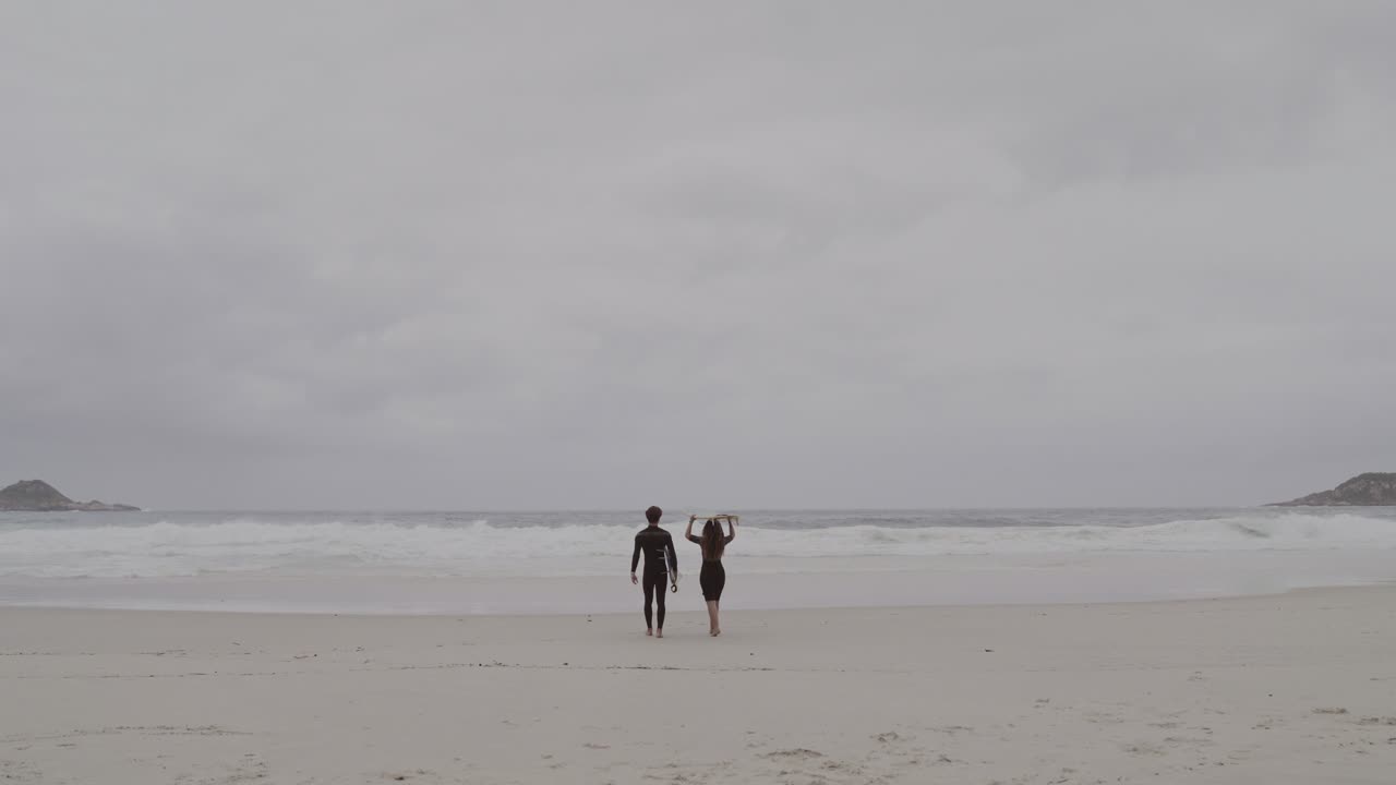 Two surfers walk on a sandy beach towards the ocean under a cloudy sky