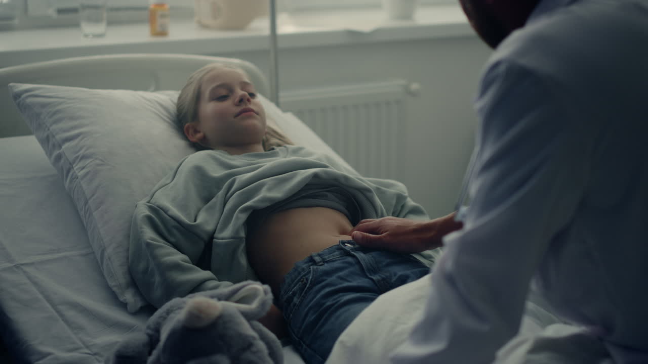Doctor palpating girl stomach ache in ward. Sick child lying in hospital bed.