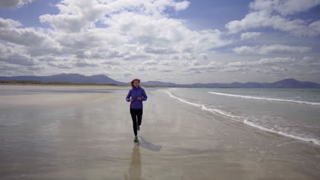 dolly shot of a girl running, jogging on the shore of a sandy beach with atlantic ocean waves on a wonderful sunny day in ireland in 4k