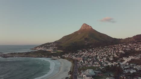 el dron vuela alto y lento sobre la playa de camps bay en ciudad del cabo, sudáfrica. en el fondo, la montaña de la cabeza de león se eleva iluminada por la puesta de sol.