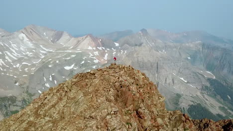 Aerial-View,-Person-With-Backpack-Standing-Alone-on-Rocky-Clifftop-With-Stunning-View-of-Mountain-Range-on-Sunny-Day