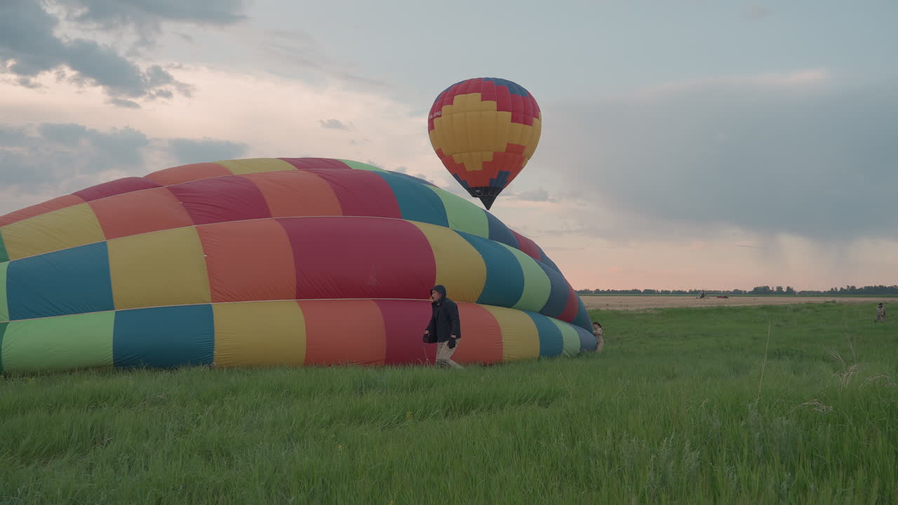 crew prepping backup hot air balloon envelope while first vibrant balloon floats above lush field at sunset capturing teamwork and scenic landscape under moody clouds ready for next launch
