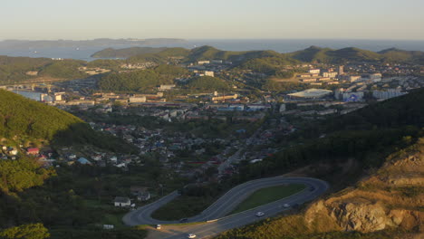 Provincial-city-with-a-serpentine-road-over-the-mountain-pass-with-moving-cars-in-the-sunset,-Russia,-Far-east