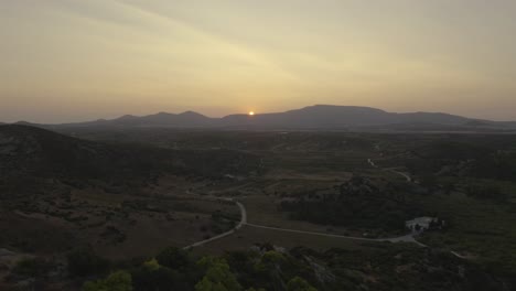 drone flying backwards with a view of a countryside rural landscape with the sun setting on top of a mountain