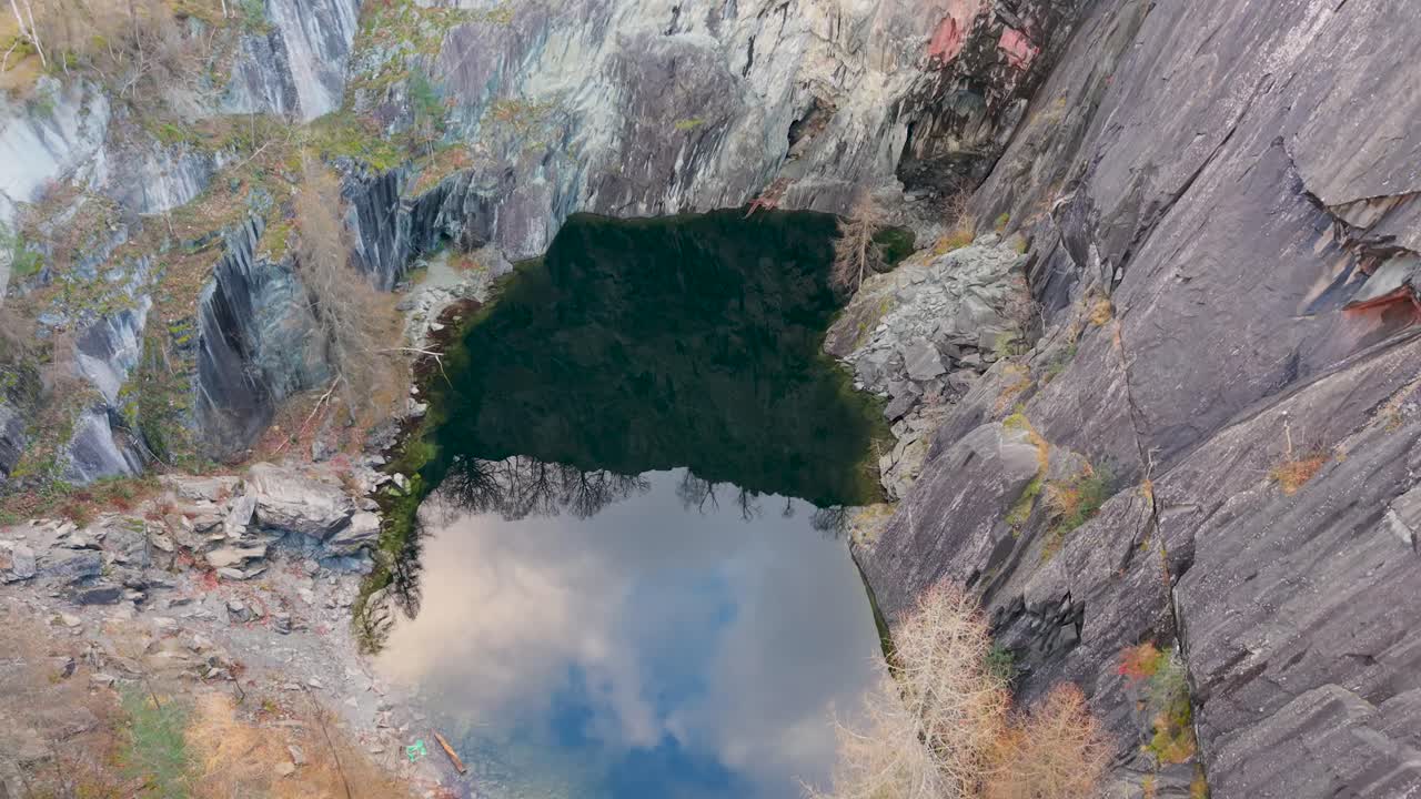 A drone faces the cave at a steep angle, gently ascending to reveal the towering slate cliffs of Hodge Close Quarry. Reflections of sky and drifting clouds shimmer across the still lake below