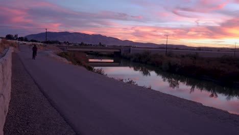 cyclist bikes down a path along a river as a brilliant sunset reflects off the water