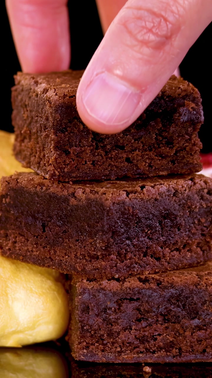 Parlak stüdyo ışığı altında, siyah bir arka plan ve yakın çekim perspektifiyle, bir el dikkatlice çikolatalı bir brownie'yi bir yığın brownie ve sarı hamur işinin üzerine yerleştiriyor.