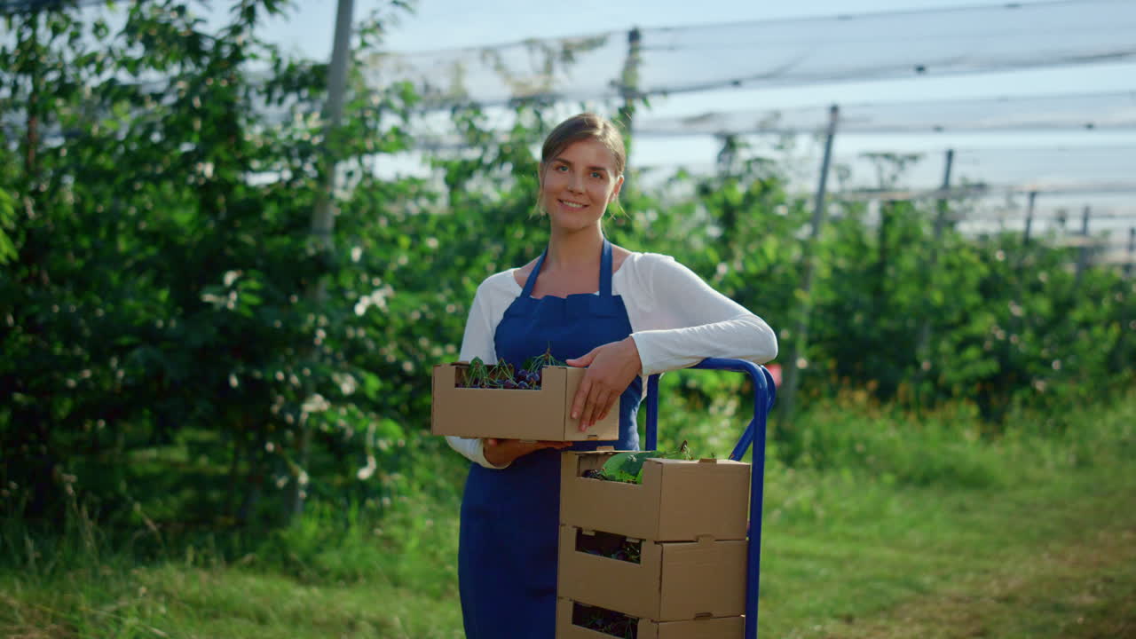 hermosa granjera buscando cámara cerca de la caja de frutas frescas en el huerto agrario.