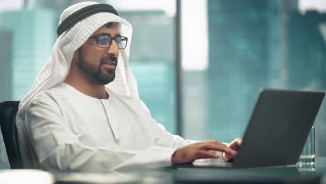 successful arab businessman in glasses and white traditional outfit sitting in office and working on laptop. business manager planning corporate strategy. saudi, emirati, arab businessman concept.