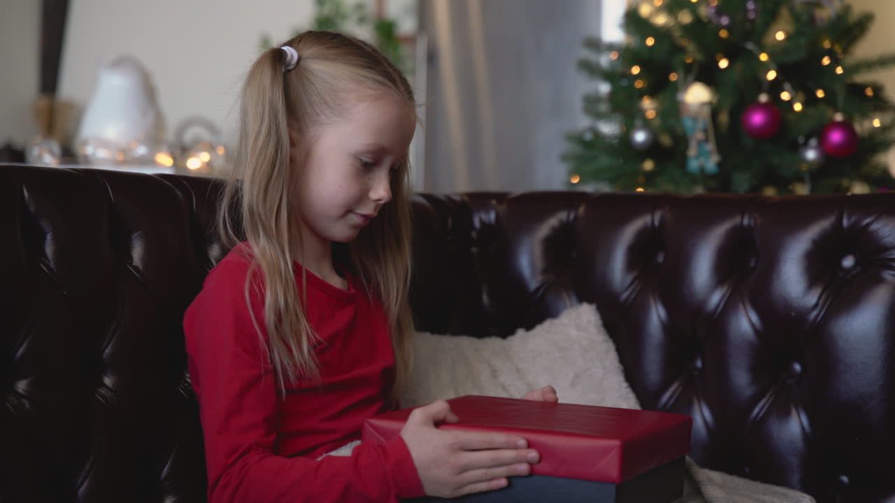 Blond pige åbner en julegave, siddende på sofaen i en stue med julepynt