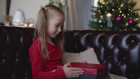 blonde girl opens a christmas present sitting on the sofa in a living room with christmas decorations