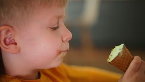 close up side view of little boy in orange shirt licking ice cream cone indoors with soft blur in background, capturing focused joyful moment of child