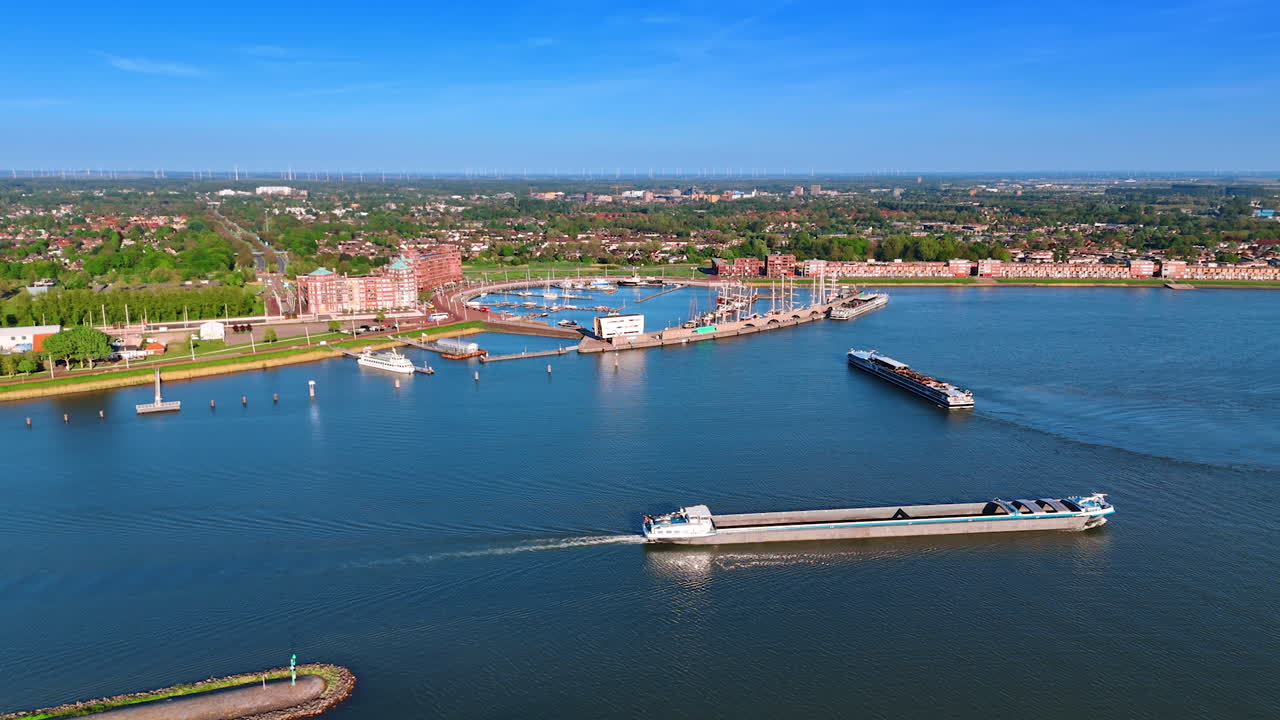 Long barges move by the peaceful waterscape on sunny daytime. Port of Lelystad, the Netherlands and cityscape in lush greenery at backdrop. Aerial view.