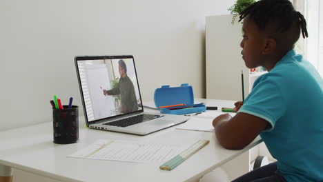 african american boy doing homework while having a video call with male teacher on laptop at home
