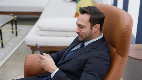 exited businessman relaxing with smartphone, texting on cellphone seated in armchair, online gaming while at office using wifi internet connection, browsing web, smiling writing message mobile phone