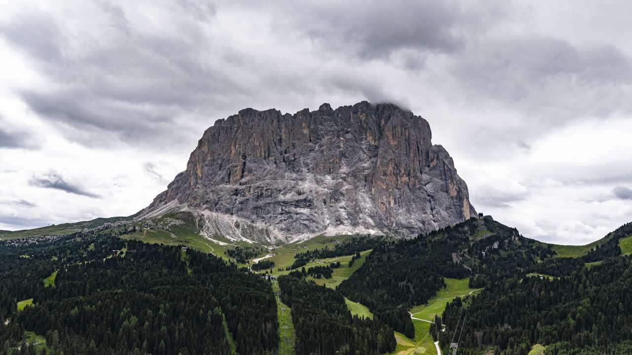 Berget Sassolungo dominerar den gröna dalen med skogar och molnig himmel i de italienska Dolomiterna.