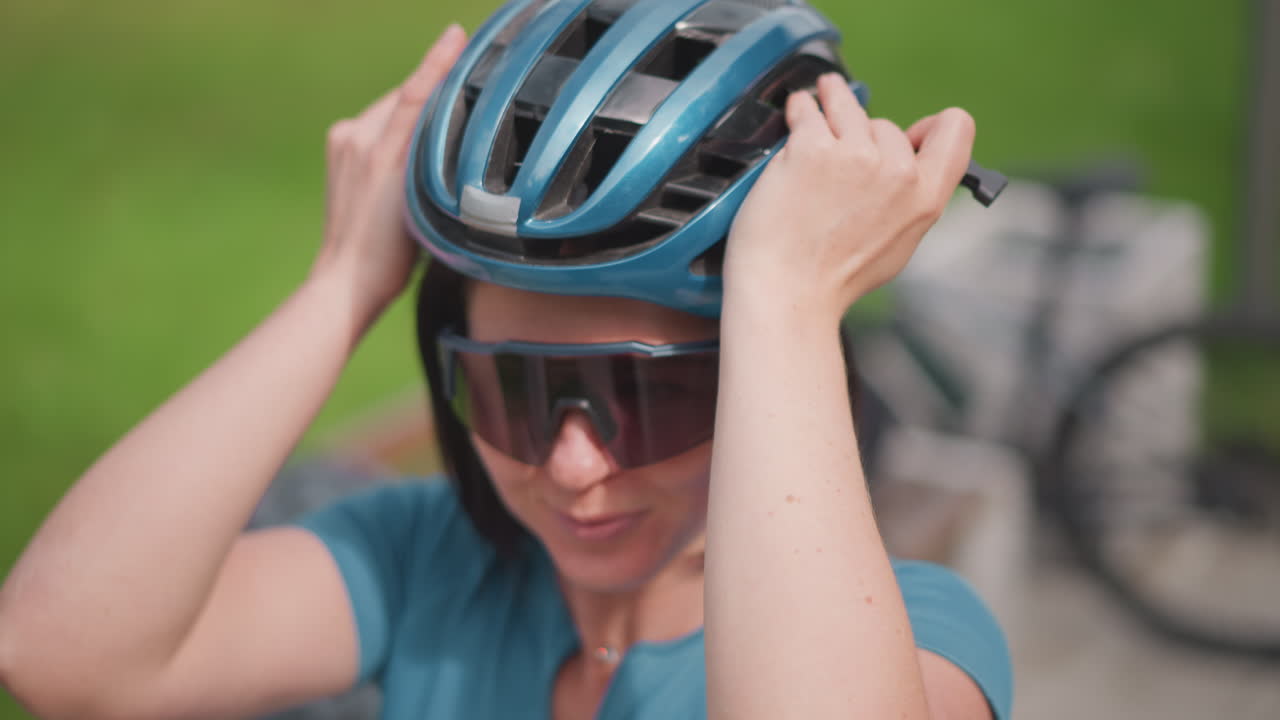 woman preparing for bike ride smiling while adjusting sunglasses and securing blue helmet on head on park bench bicycle parked in background sunny green setting closeup captures safety routine