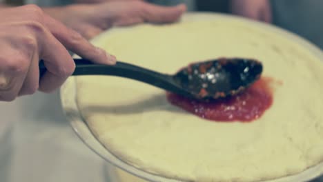 italian chef taking fresh tomato sauce from a bowl and spreading it in circle motions on the pizza dough