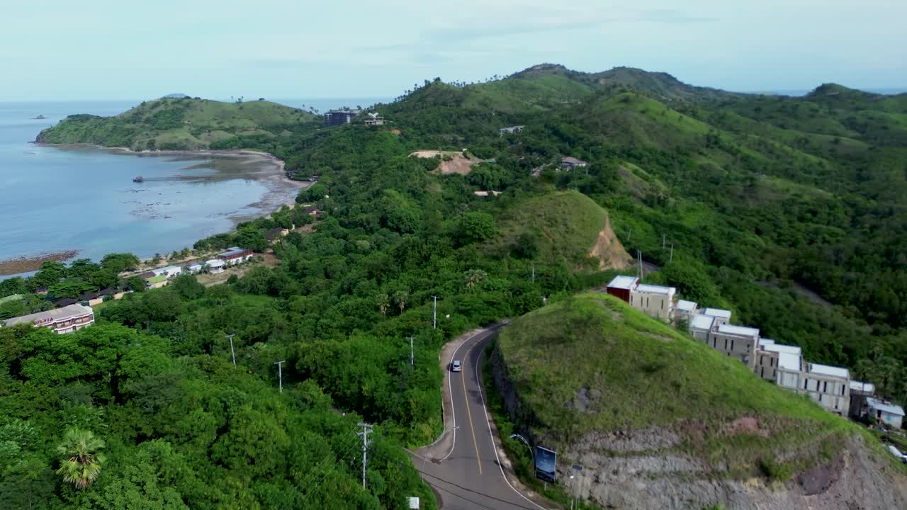 Una ripresa aerea cinematografica di una strada tortuosa che attraversa lussureggianti colline tropicali lungo una costa serena. Circondata da vegetazione, vista sull'oceano e una delicata luce diurna, questa scena evoca sensazioni di libertà.