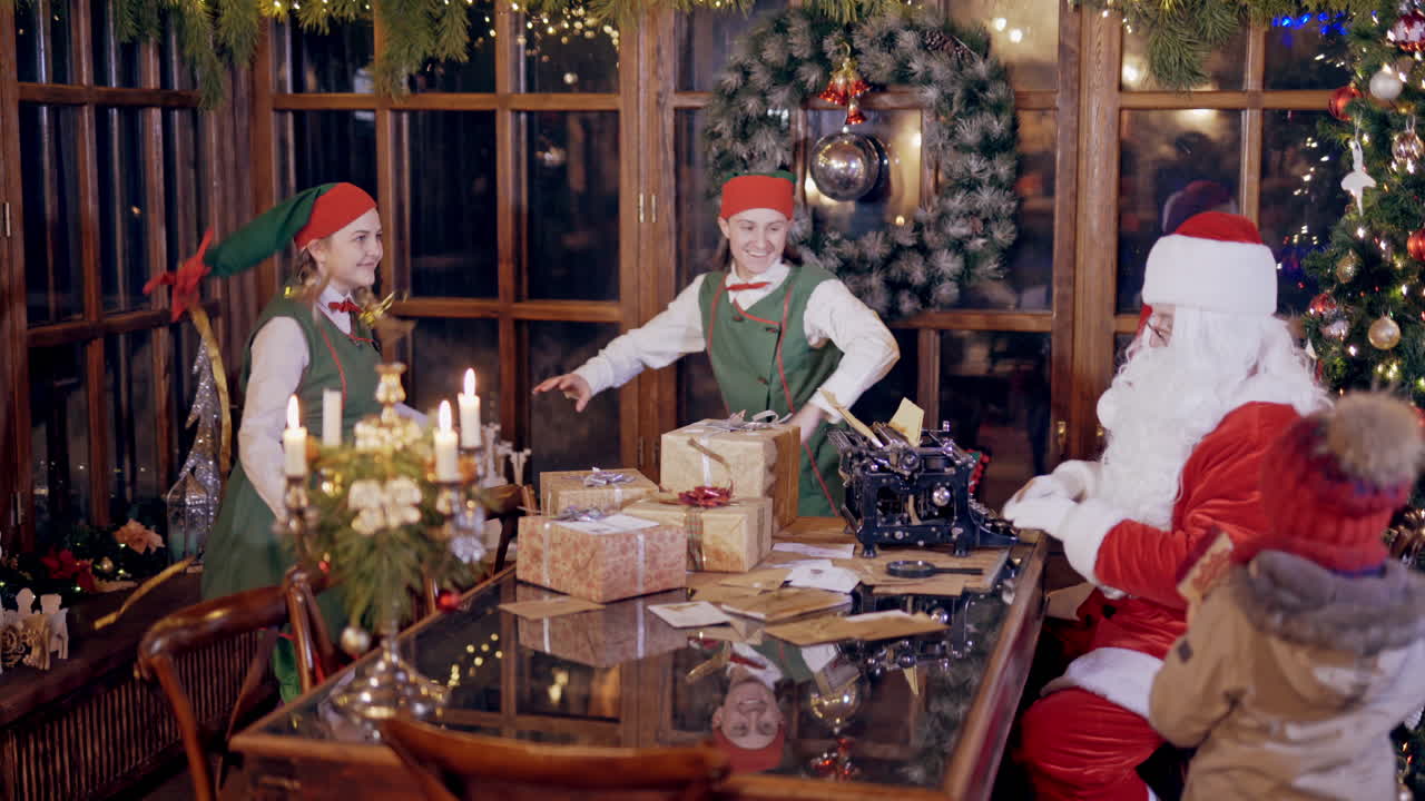 Joyful elves and Santa in festive costumes. Santa is busy at typewriter while two elves dancing inside Santa's residence. Christmas atmosphere.