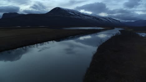 Flying-Above-River-With-Boat-Cruising-On-A-Cloudy-Sunset-With-View-Of-Kebnekaise-Mountain-In-Swedish-Lapland