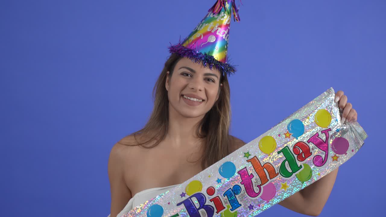 primer plano de una mujer joven posando con un letrero de feliz cumpleaños y un sombrero de fiesta, aislada en un fondo de estudio azul