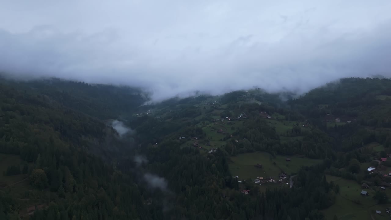 Panoramic drone view of Mătișești village nestled in a misty valley of the Apuseni Mountains in Romania, with clouds drifting between forested slopes and scattered rural homes after sunset