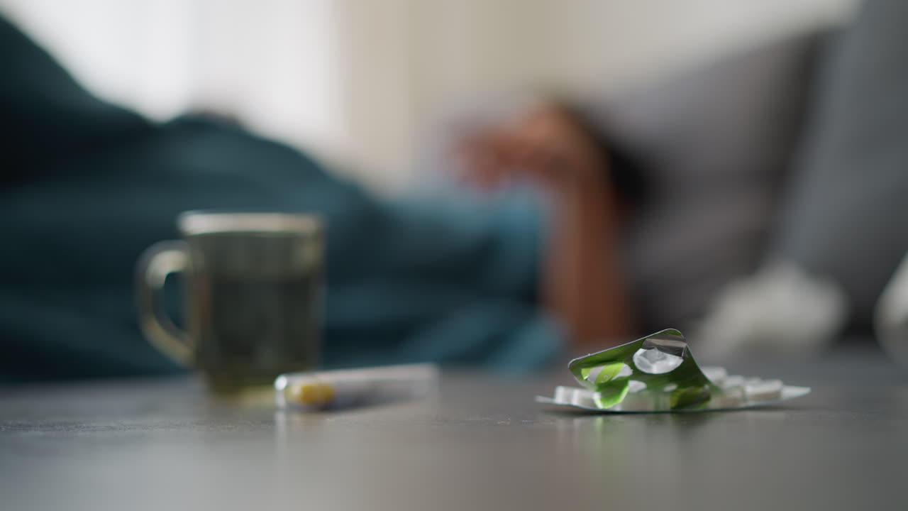 Close-up of medication and cup on table with blurred view of person adjusting on bed, suggestive of recovery, rest, and care during illness with soothing indoor setting