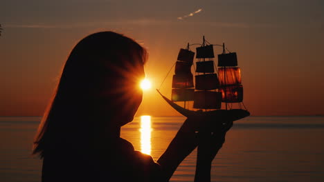 A-Woman-Stands-By-The-Sea-At-Sunset-Holds-In-Her-Hands-A-Model-Of-A-Sailing-Ship-Hope-And-Travel-Con