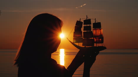 una mujer se encuentra junto al mar al atardecer sostiene en sus manos un modelo de un velero esperanza y viaje con