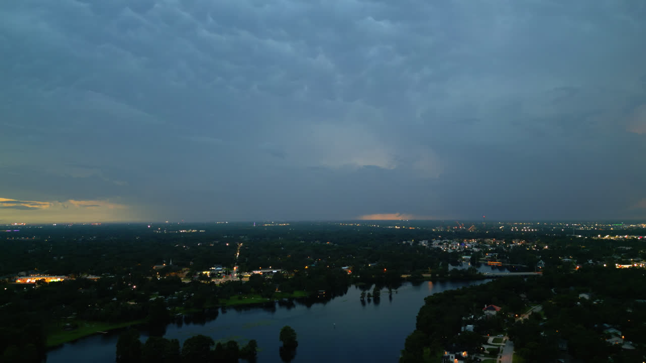 Aerial slow tracking shot over a river reflecting the evening twilight sky as lightning flashes in the clouds and the city skyline with soft ambient light being turned on