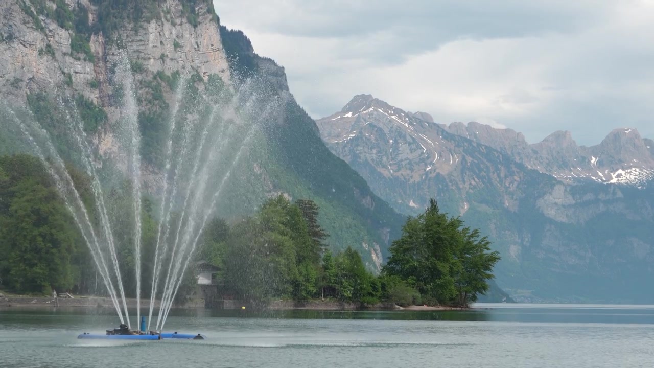Tall fountain jets spraying water across calm Walensee lake with snowcapped Swiss Alps and forested cliffs behind in Weesen Switzerland