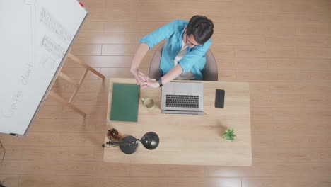 top view zoom out shot of a man stretching while sitting at the wooden desk besides a whiteboard typing on a laptop next to the smartphone at home