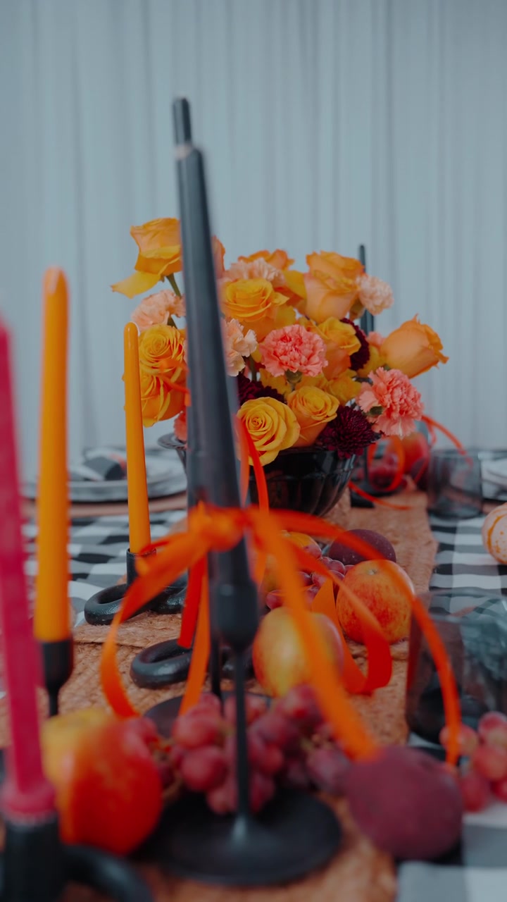 Elegante allestimento tavola a tema autunnale con vivaci candele arancioni, centrotavola floreale e frutta decorativa su una tovaglia fantasia