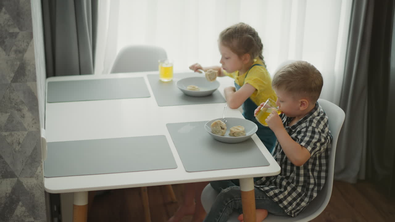 little girl wearing yellow top and overalls eating meal from bowl while watching young boy in checkered shirt drinking fruit juice at modern dining table with sunlight through white curtains