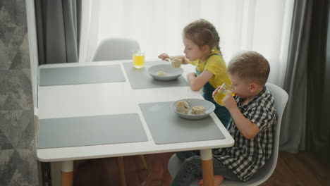 little girl wearing yellow top and overalls eating meal from bowl while watching young boy in checkered shirt drinking fruit juice at modern dining table with sunlight through white curtains