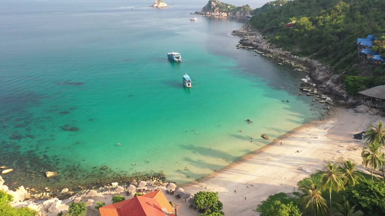 Video aereo con drone di una baia esotica in un'isola paradisiaca tropicale, caratterizzata da formazioni calcaree, alberi e resort, con una spiaggia di sabbia bianca cristallina color smeraldo, visitata da barche a vela, in Thailandia