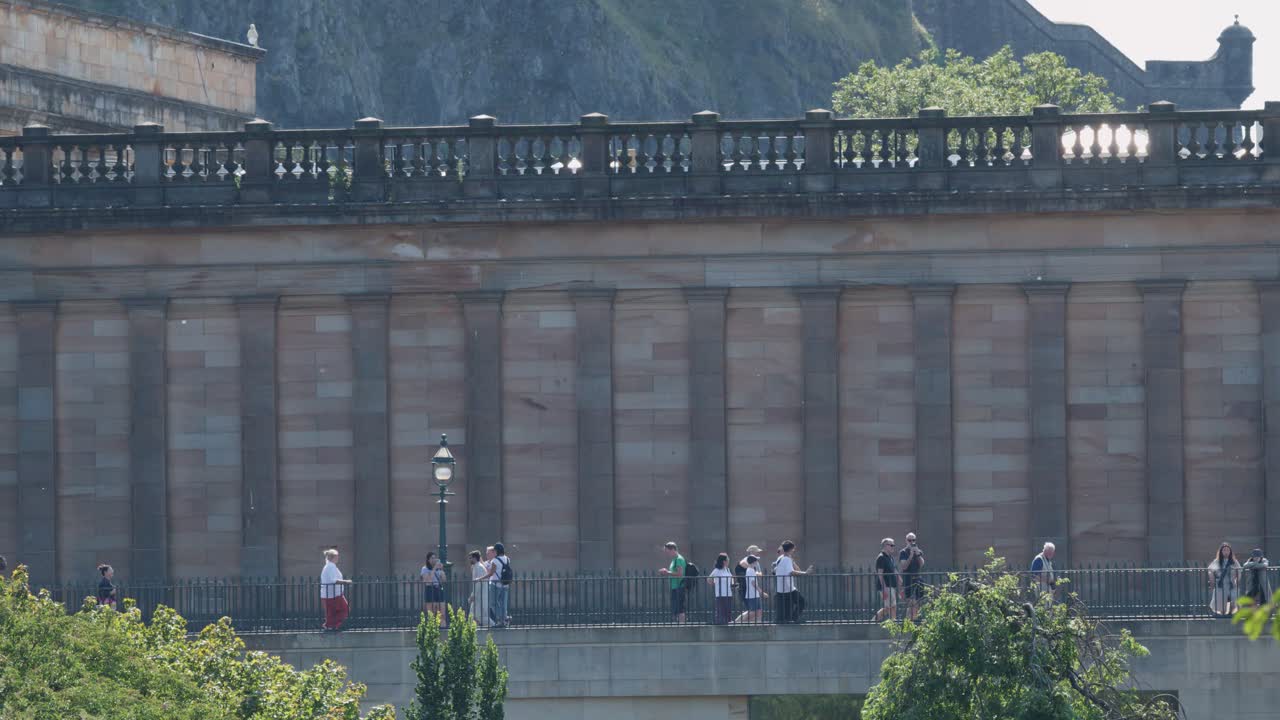 Groups of people walk along gallery facade, midday sunlight, static wide shot, Edinburgh urban setting