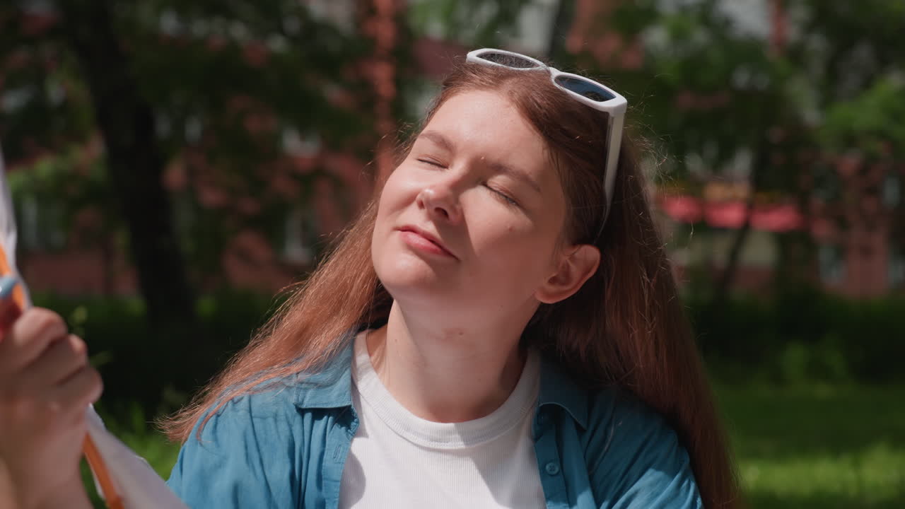 Female student seated outdoors under bright sunlight, fanning herself gently with embroidered fabric hoop, enjoying warm summer day near residential building surrounded by trees