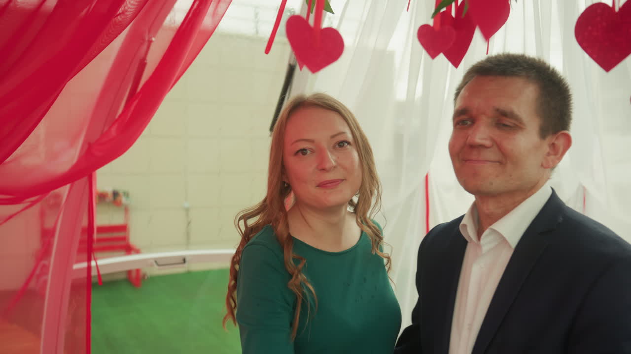 Ett par i ett rött dekorerat bås delar champagne och varma leenden under ett Alla hjärtans dag-date. Kvinnan hälsar och skakar hand och poserar sedan nära mannen under hängande hjärtdekorationer och mysiga draperade gardiner.