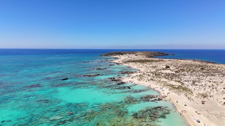 La spiaggia di Elafonissi presenta acqua turchese poco profonda, sabbia chiara e formazioni costiere rocciose a Kissamos, Chania, Creta, Grecia, durante un sorvolo in avanti con drone.